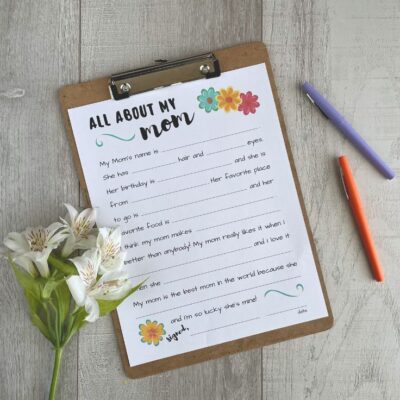 All About My Mom Printable interview worksheet in a frame with pens & flowers on a wood backdrop.