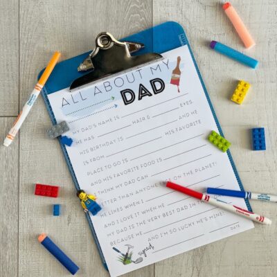 Blue clipboard with a fill in the blank Father's Day worksheet attached, surrounded by legos, darts & markers.