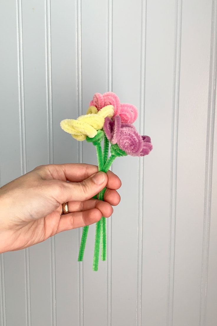 A hand holding a bouquet of flowers made from yellow, pink, purple, and green chenille stems.