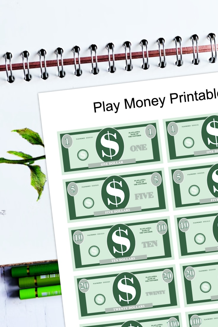 Preview of printable money PDF page on top of a white art spiral notebook and 3 green colored pencils on bottom. 