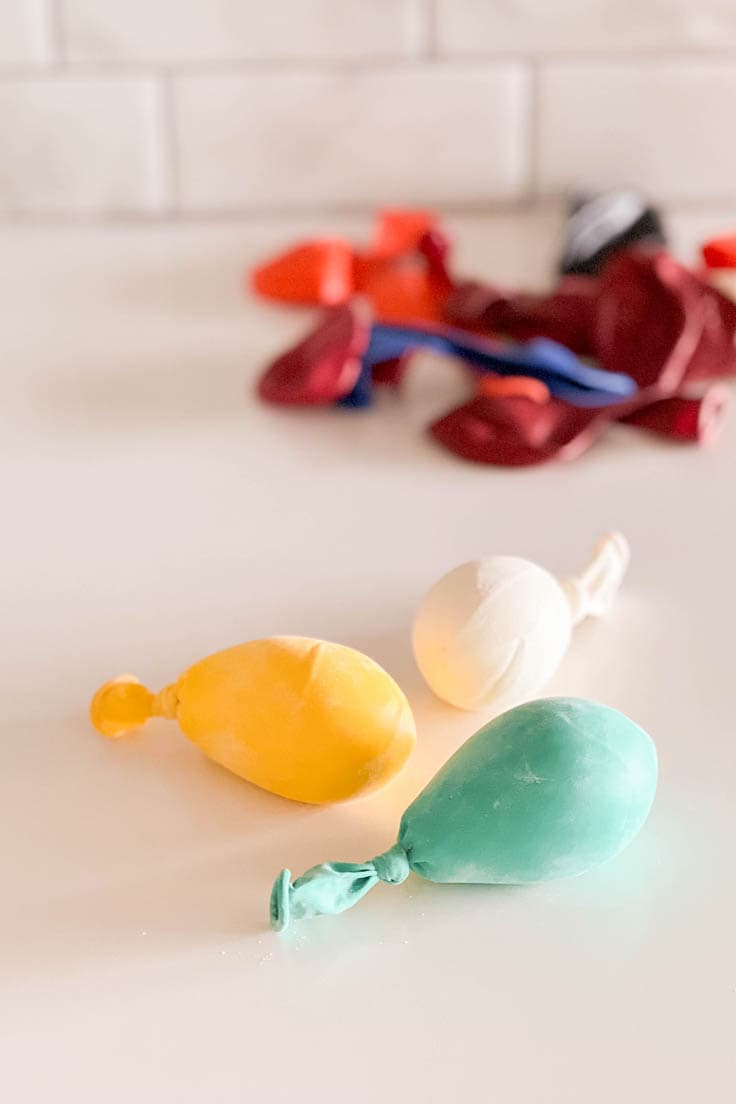 Three homemade mini balloon stress balls, one yellow, one white, and one aqua sitting on a white table with deflated balloons in the background
