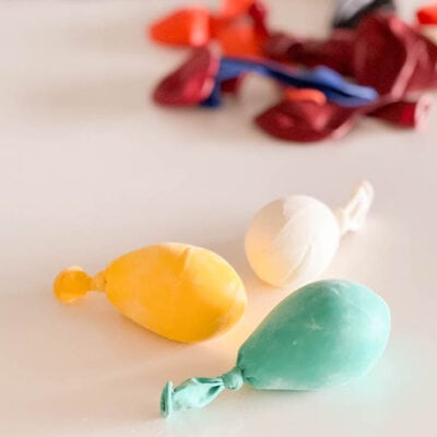 Three homemade mini balloon stress balls, one yellow, one white, and one aqua sitting on a white table with deflated balloons in the background