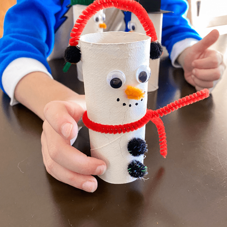 child holding a toilet paper snowman with ear muffs and a red scarf