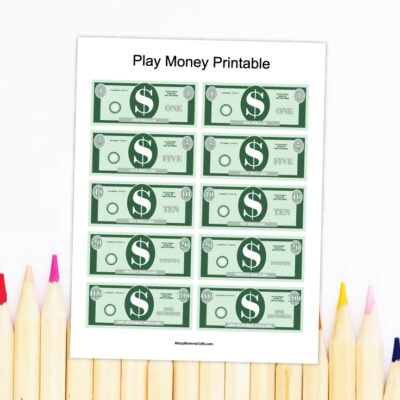 Preview of printable play money PDF on top of white background with wooden colored pencils on bottom.