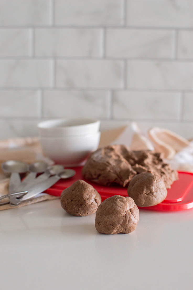 Homemade cinnamon playdough divided into 3 small spheres and the remaining playdough sitting on a red cutting board in the background. Image also contains measuring spoons, a plaid napkin, and a small bowl of ground cinnamon.
