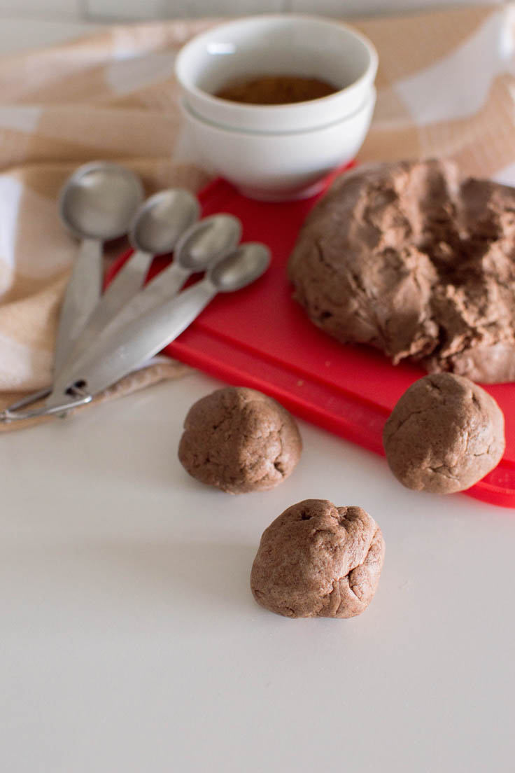 Homemade cinnamon playdough divided into 3 small spheres and the remaining playdough sitting on a red cutting board in the background. Image also contains measuring spoons, a plaid napkin, and a small bowl of ground cinnamon.