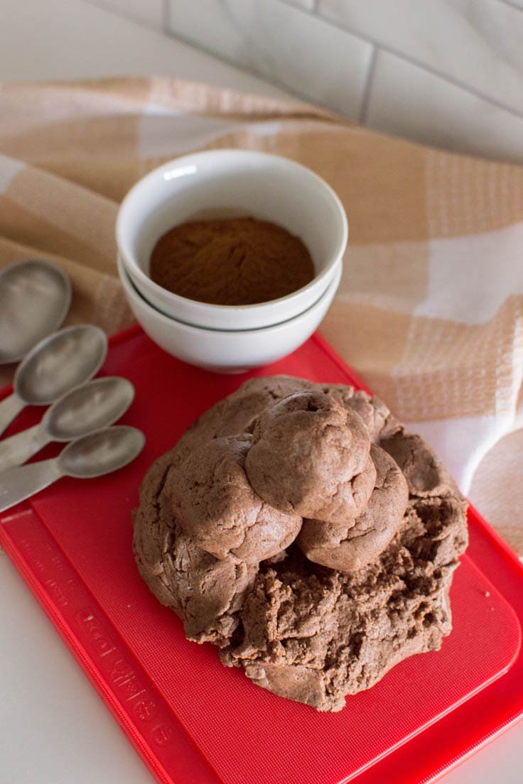 Cinnamon playdough divided into 3 small spheres and sitting on top of a larger mound of playdough, sitting on a cutting board in the background. Image also contains measuring spoons, a plaid napkin, and a small bowl of ground cinnamon.