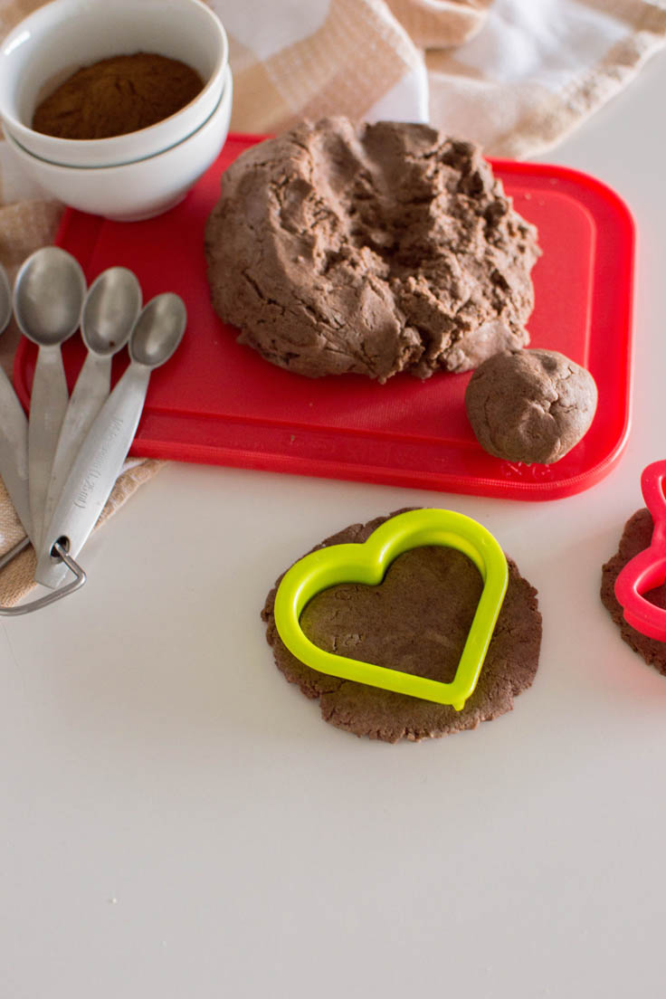 A green heart-shaped cookie cutter cutting into a small portion of no-cook cinnamon playdough. Surrounded by more playdough, a red cutting board, measuring spoons.