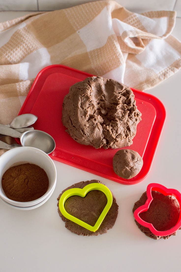 Cookie cutters surrounding cinnamon playdough, sitting on a red cutting board and surrounded by a plaid napkin and two small white bowls, stacked on top of each other.