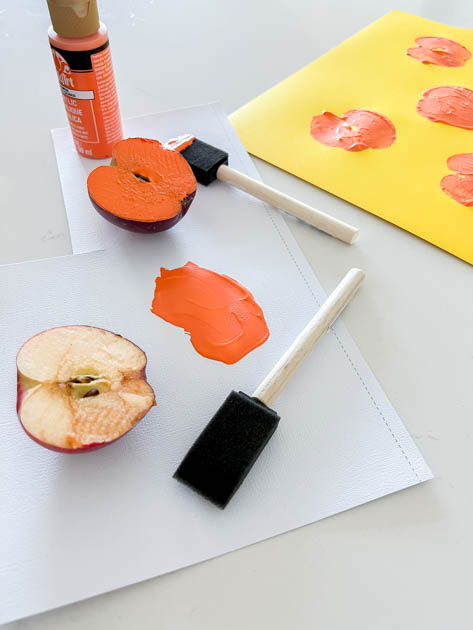 Foam paint brush, apple halves, and orange paint sitting on a white surface