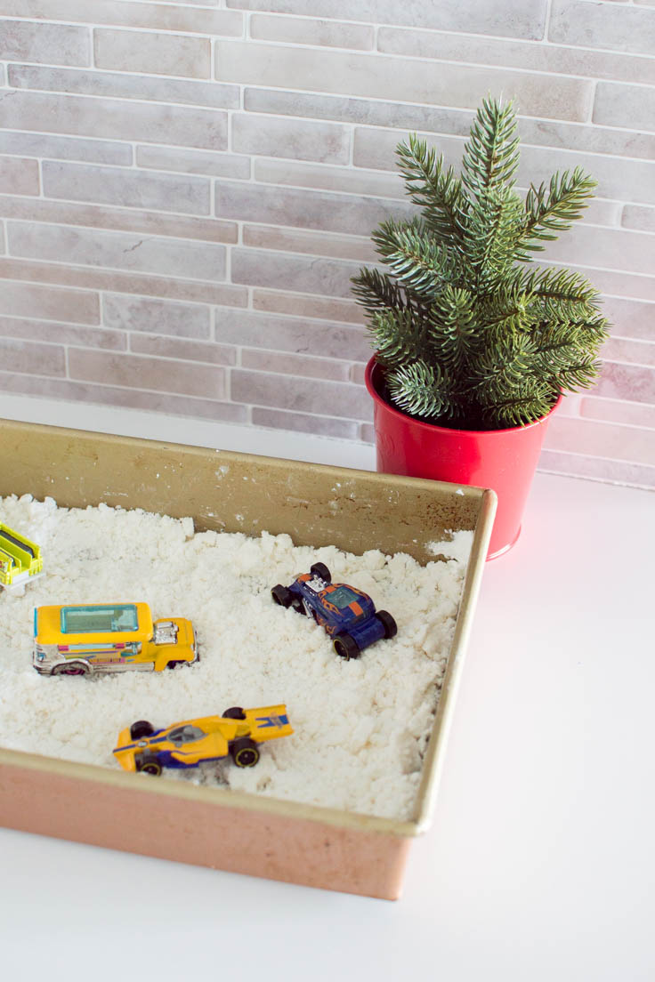 A portion of a baking pan shown, it is filled with pretend snow and toy cars. A miniature Christmas tree in a red vase sits in the background.