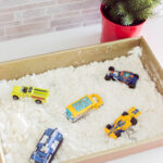 Aerial view of DIY Fake Snow for Sensory bins, sitting in a baking pan with some Hot Wheels cars