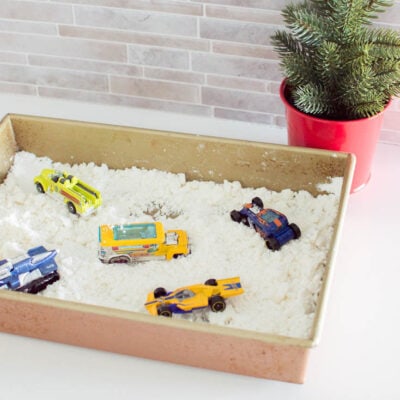A baking pan filled with DIY fake snow and toy cars, with a faux mini Christmas tree in the background