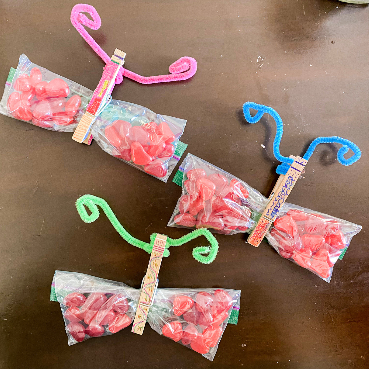 3 jelly bean butterflies: baggie with heart shaped jelly beans and a clothes pin in the middle with pipe cleaner antenna, top has pink pipe cleaner, middle has blue, and bottom has green