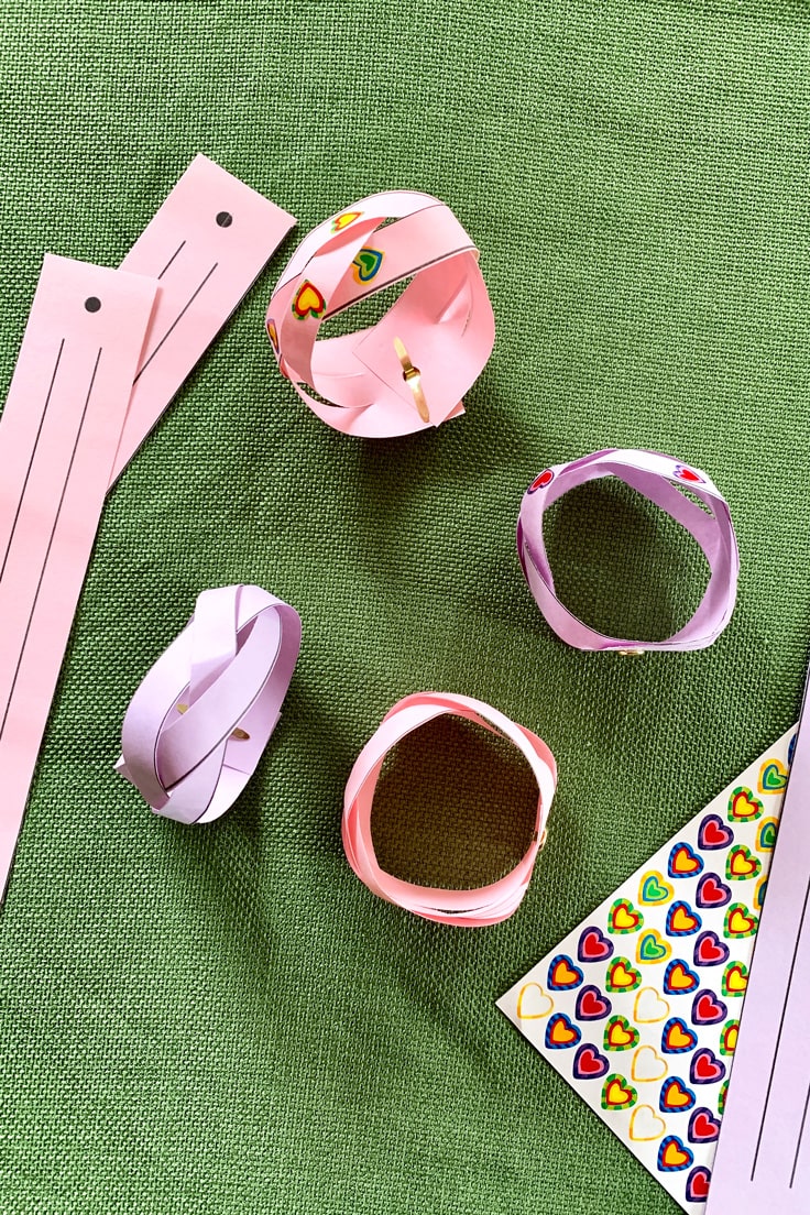 Braided paper bracelets in pink and purple with heart stickers on a green tablecloth.