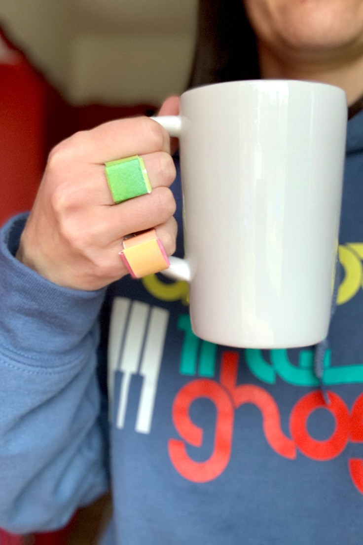 Person holding a coffee mug and wearing green and orange paper rings on two fingers.