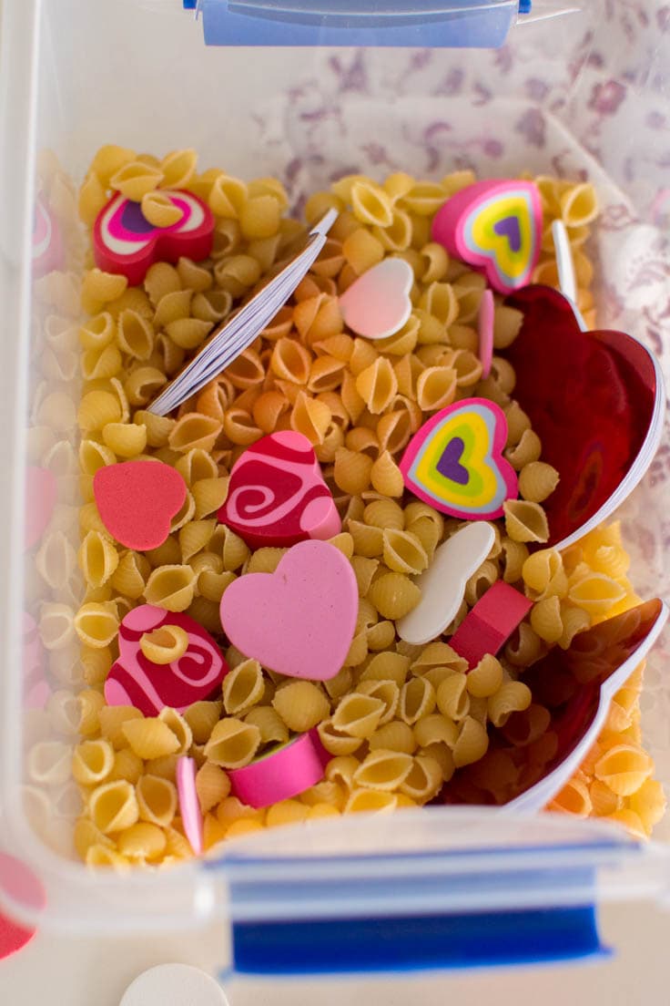 Aerial view of heart-shaped items in a bin full of pasta to make a Valentine's Day sensory bin