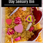 Aerial view of heart-shaped items in a bin full of pasta to make a Valentine's Day sensory bin