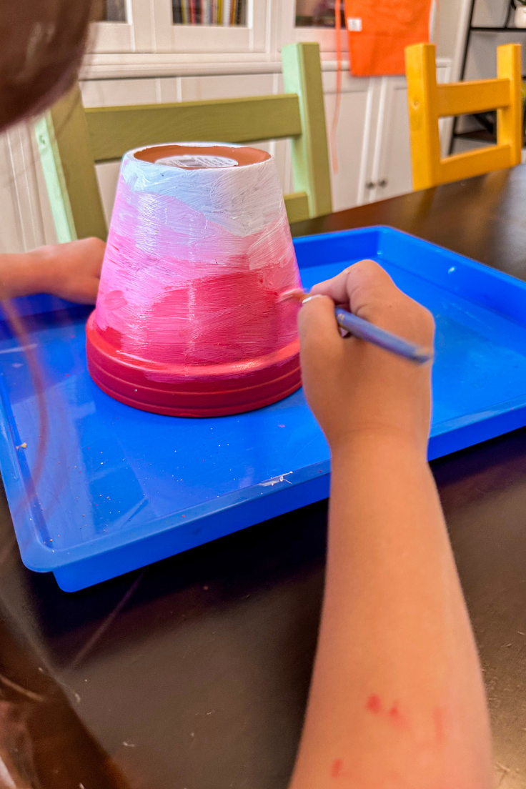 child mixing the red and white paint together on the pot to create a pink paint and ombre effect