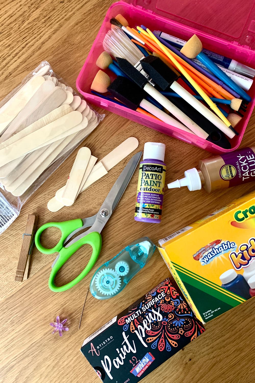 Scattered craft supplies including paintbrushes, popsicle sticks, scissors, paint, craft glue, glue dots, a clothespin, a whirly bead, and a head pin.