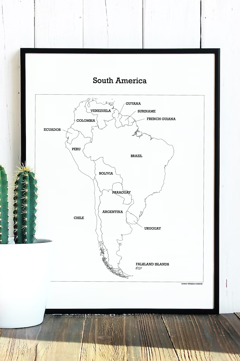 Preview of map of South America in wall art blank frame leaning on white wooden plank wall with cactus potted plant on left side. 