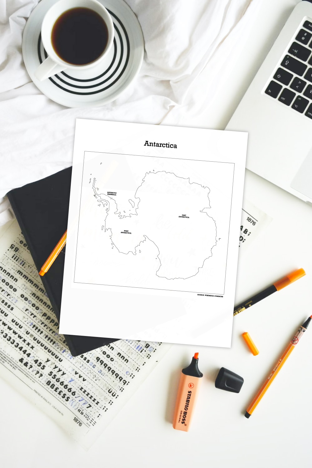 Preview of printable map of Antarctica on white desk with mug of coffe above, corner of laptop computer keyboard in upper right and notebook and orange highlighter and writing markers below. 