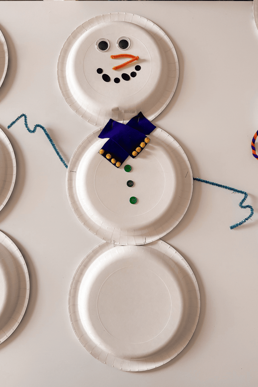 snowman made from paper plates with orange pipe cleaner nose and a blue scarf 