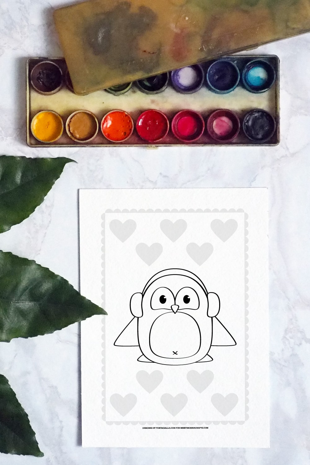 Preview of a penguin coloring page on white marble countertop with leaves on the left border and  vintage watercolor palette and paints on the top.