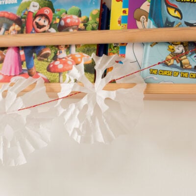 A garland made up of red string and coffee filter snowflakes hanging off a wall bookshelf