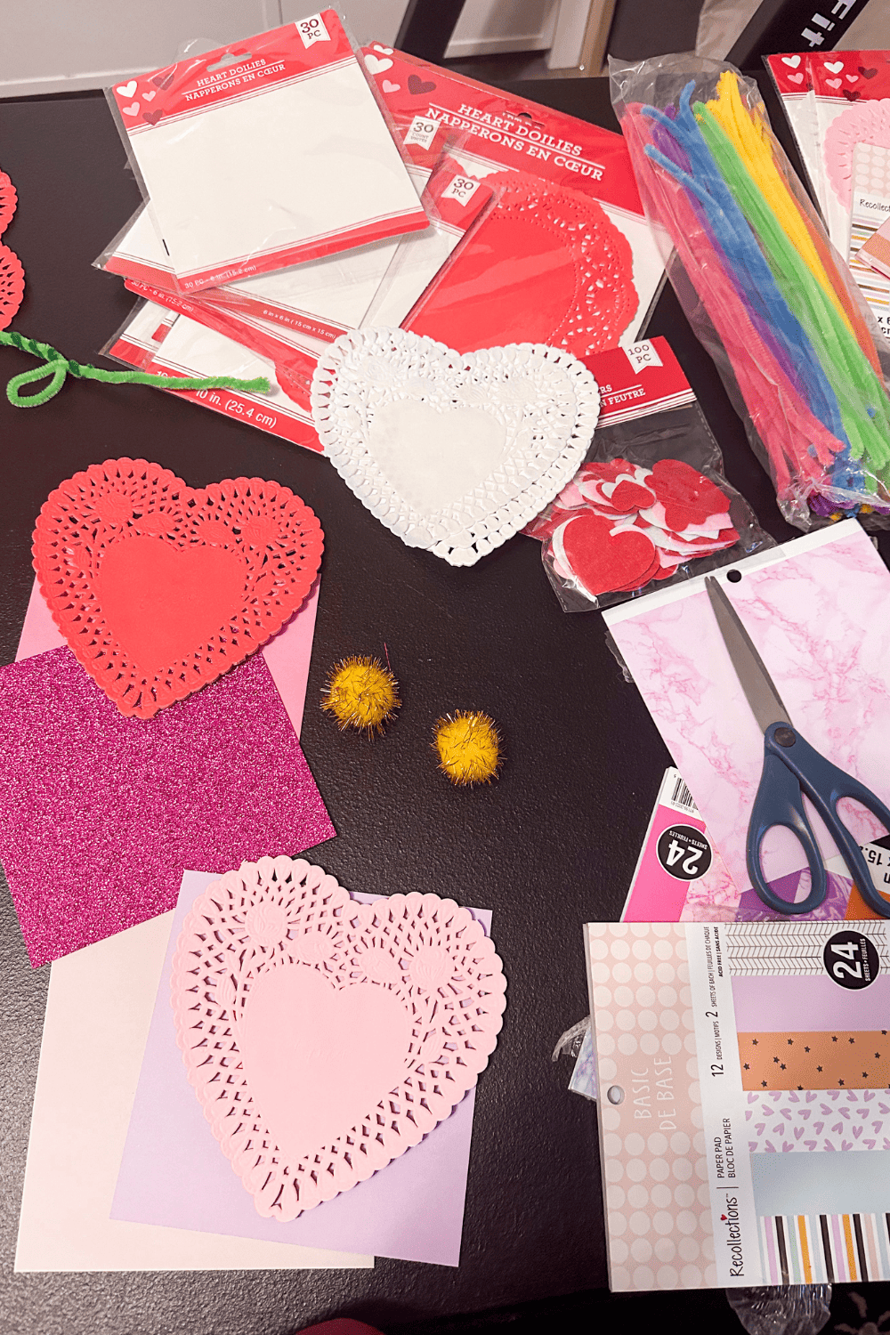 craft supplies on a table, heart doilies, construction paper, scissors, pom poms, pipe cleaners, felt hearts