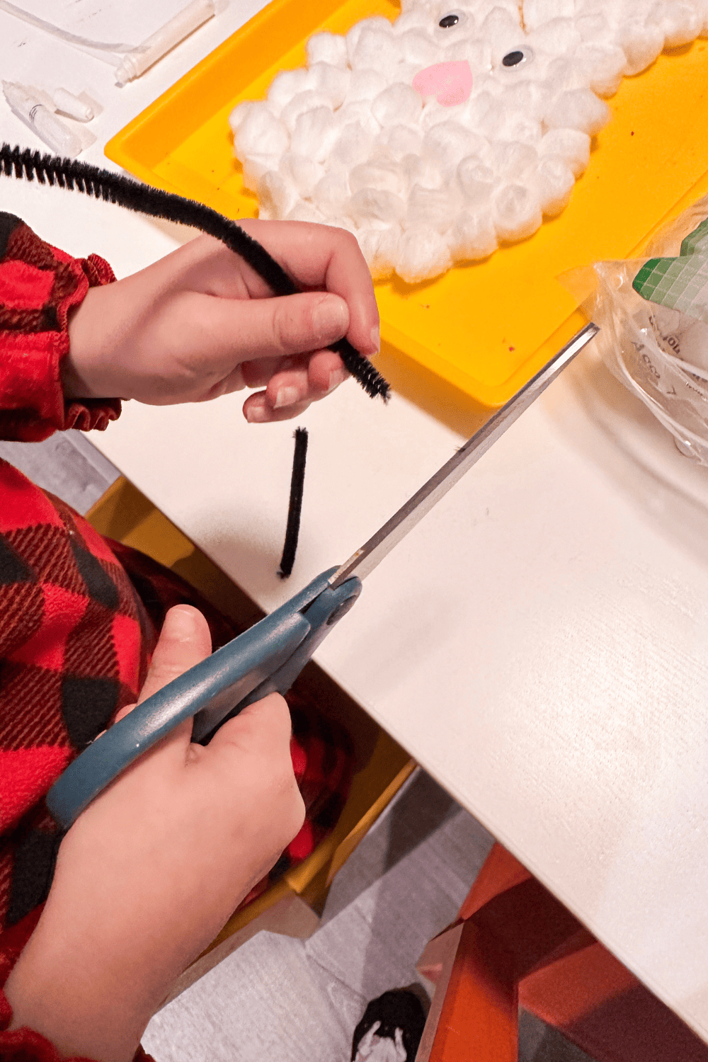 small hands holding scissors in one hand and black pipe cleaner in the other, cotton balls in the background