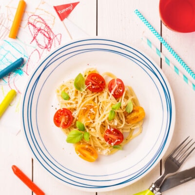 A plate of pasta with tomatoes on a kids' table with crayons and paper.