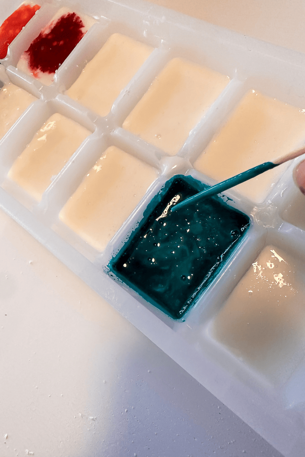 A toothpick covered in homemade watercolor paint being held slightly above a blue homemade watercolor paint in an ice cube tray